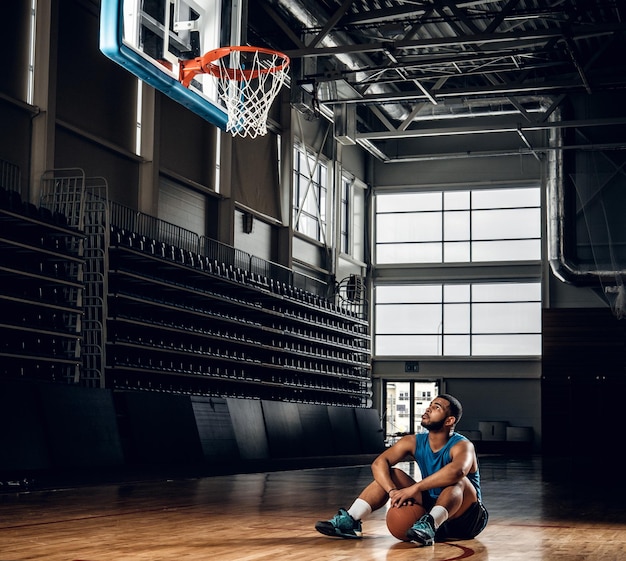 Portrait of Black basketball player sits on a floor under  a hoop in a basketball hall.