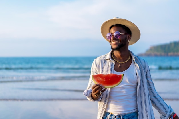 Портрет чернокожего этнического мужчины наслаждается летними каникулами на пляже, поедая арбуз