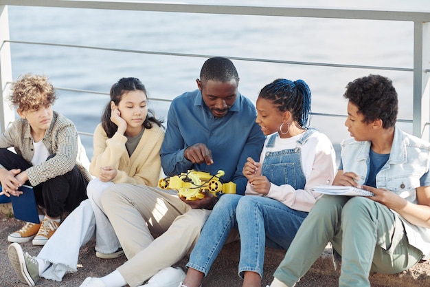 Portrait of black male teacher showing robot model to kids outdoors during engineering class