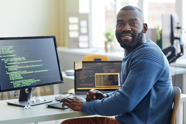 Portrait of black man as computer programmer looking at camera while writing code at office workplac