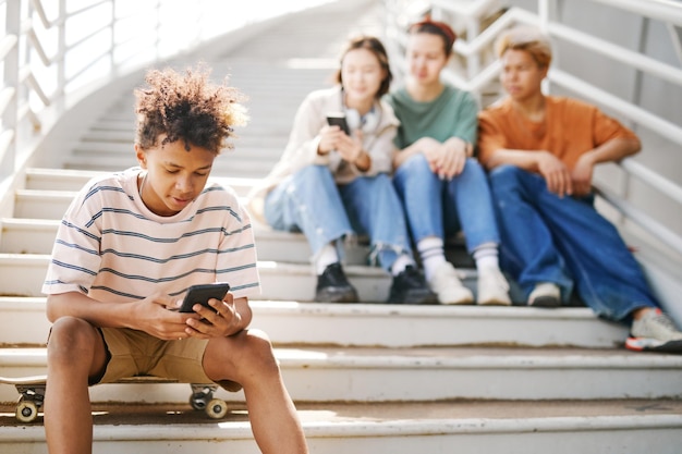 Photo portrait of black teenage boy using smartphone outdoors while sitting on metal stairs with group of