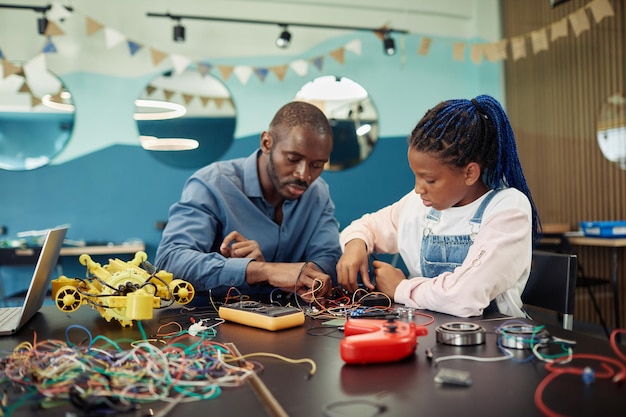 Portrait of black young girl building robots with male teacher helping during engineering class at s