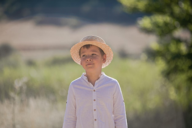 Portrait of the blode hair boy in straw hat with blurred summer greenery background