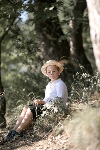 Portrait of the blode hair boy in straw hat with blurred summer greenery background