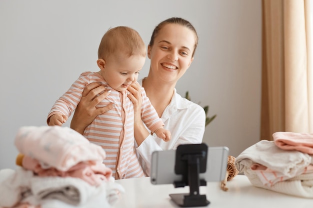 Photo portrait of blogger woman sitting at table with toddler kid and recording video for her vlog, making content with her daughter, showing kid to followers and smiling.