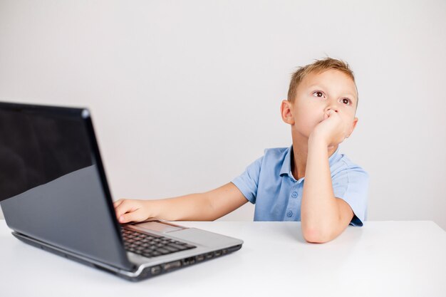 portrait of blond boy sitting at desk and looking up pensively while using laptop