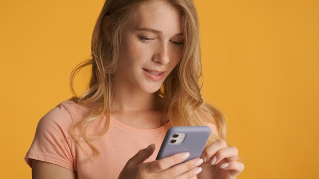 Portrait of blond girl texting with friend in online chat on smartphone over yellow background Surfes social media