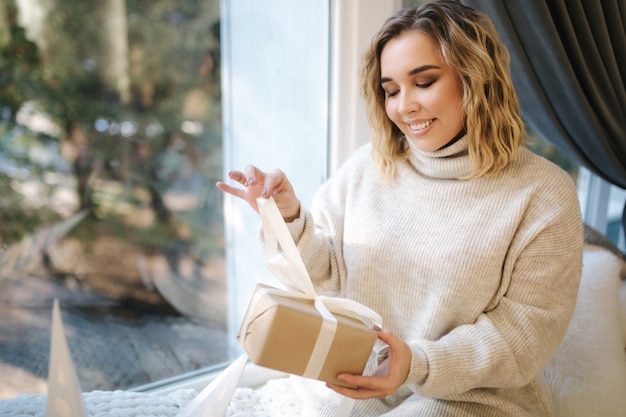 Portrait of blond hair woman in hoodie open present on Chrisrmas time. Elegant woman stand by big