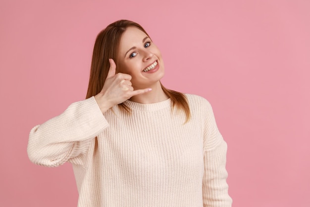 Portrait of blond woman making telephone gesture near head and winking playfully, waiting for call, wants to talk on phone, wearing white sweater. Indoor studio shot isolated on pink background.