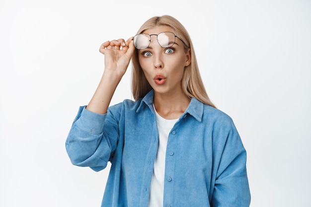 Portrait of blond woman taking off her glasses, widen eyes and staring impressed, surprised to see something, standing on white