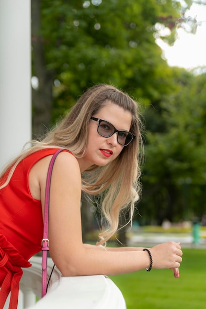 Portrait of blonde attractive young lady in elegant red dress and black sunglasses on park background Vertical frame