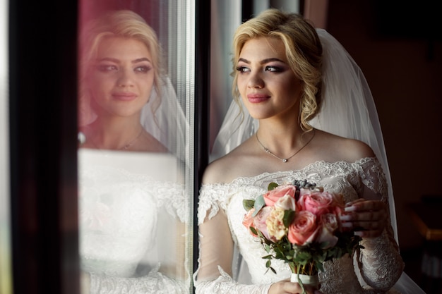 Портрет белокурой невесты с букетом розовых роз крытый. , молодая улыбающаяся невеста с красивым вырезом в роскошном платье около окна. день свадьбы.