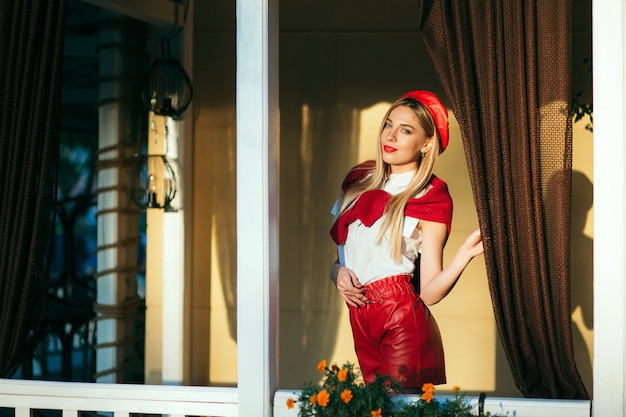 Portrait of blonde female in red beret and leather red shorts posing outdoor. Red glasses and scarf.