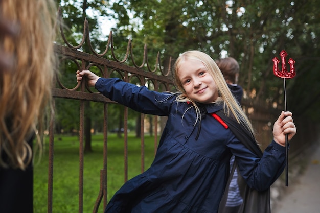 Portrait of blonde girl dressed in Halloween costume holding onto fence while playing with friends outdoors, copy space