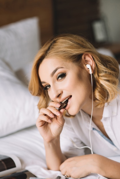 Портрет блондинка в первой половине дня, который слушает музыку через наушники в белой кровати.