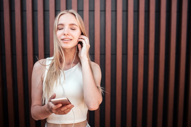 Portrait of blonde girl standing at wall and listening to music. She is keeping her eyes closed. Girl is enjoying the moment.