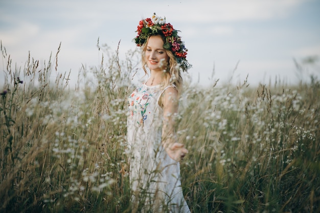 Foto ritratto della ragazza bionda con gli occhi azzurri con una corona di fiori sulla sua testa che cammina nel campo con i fiori bianchi