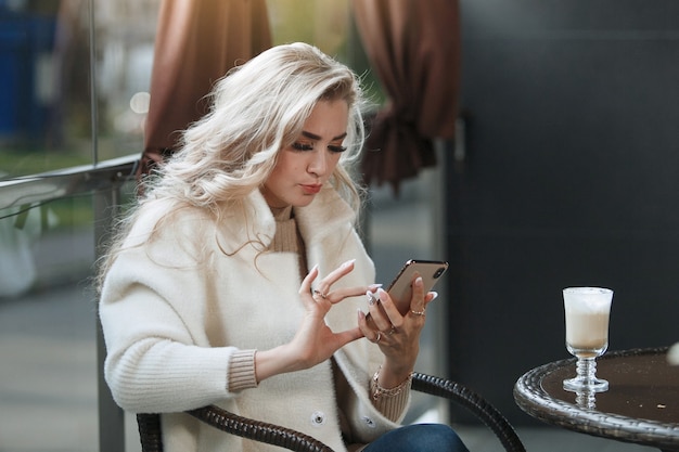 Portrait of a blonde lady at a table in a cafe looking at a smartphone, long hair, beige coat. Chatting in social networks, reading the news.