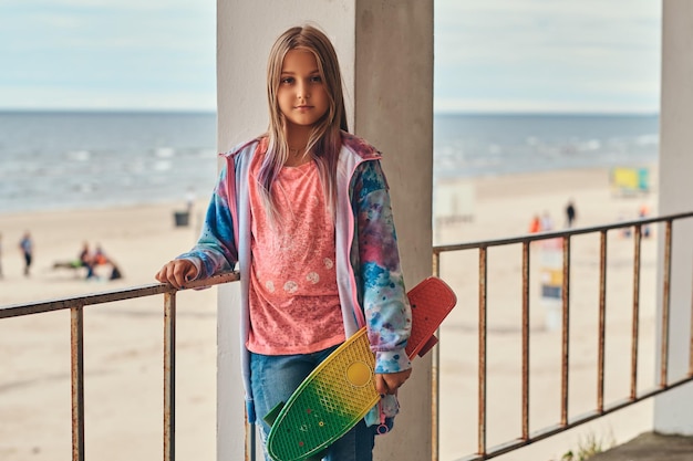 Portrait of a blonde schoolgirl skater posing with a skateboard while leaning on a guardrail against a sea coast.