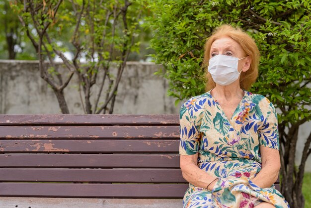 Portrait of blonde senior woman with mask thinking while sitting with distance on park bench