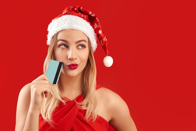 Portrait of blonde shopping girl with pensive face in Santa Claus Christmas hat with credit card thinks what gifts to buy at New Year in online store isolated on red