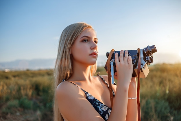 Ritratto della donna bionda in vestito dalla stampa floreale con la videocamera d'annata nel campo dell'uva durante il tramonto