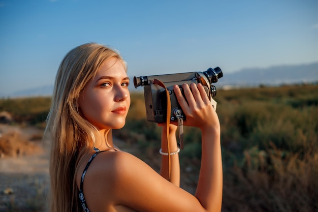 Portrait of blonde woman in floral print dress with vintage video camera in grape field during sunset