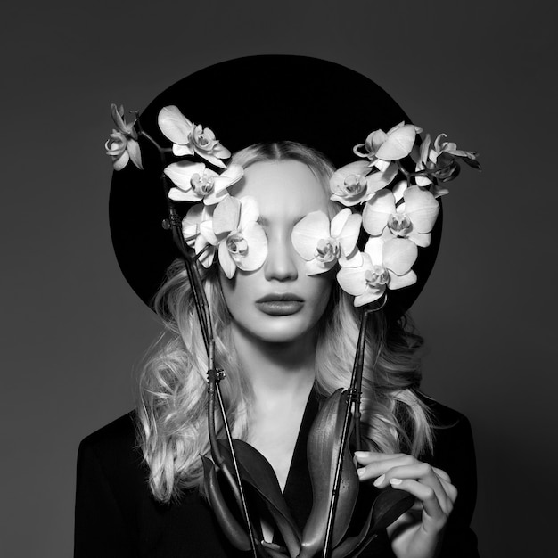 Photo portrait of a blonde woman in a large round black hat, an orchid flower in her hands