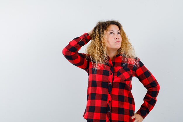 Portrait of blonde woman with curly hair keeping hand behind head, looking upward in checked shirt and looking thoughtful front view
