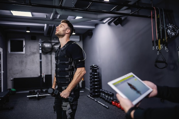 체육관 한가운데 서 있는 검은색 EMS 훈련복을 입은 남자의 신체 부위 초상화