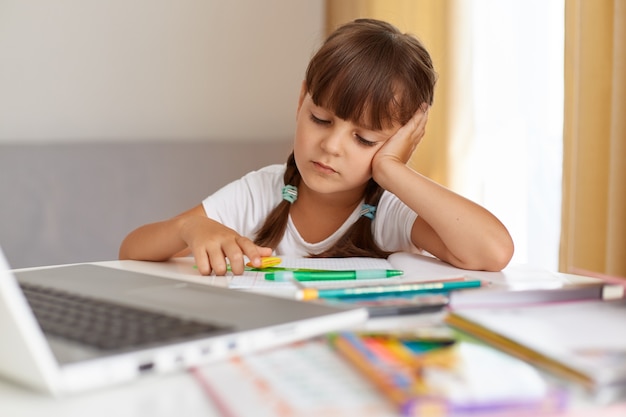 Портрет скучающей школьницы в белой футболке, сидящей за столом перед ноутбуком, с грустным выражением лица, уставшей делать домашнее задание, дистанционное обучение.