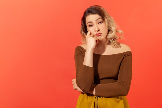 Portrait of bored young blonde woman looking into the camera with an incredulous and disappointed emotions on her face and hand near her head. Indoor studio shot isolated on red background