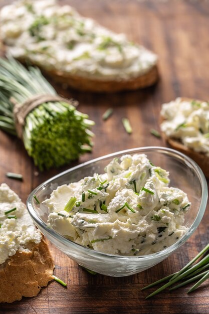 Foto ritratto di una ciotola di crema di formaggio fatta in casa da spalmare con erba cipollina tritata circondata da fette di pane con diffusione e un mazzetto di erba cipollina appena tagliata.