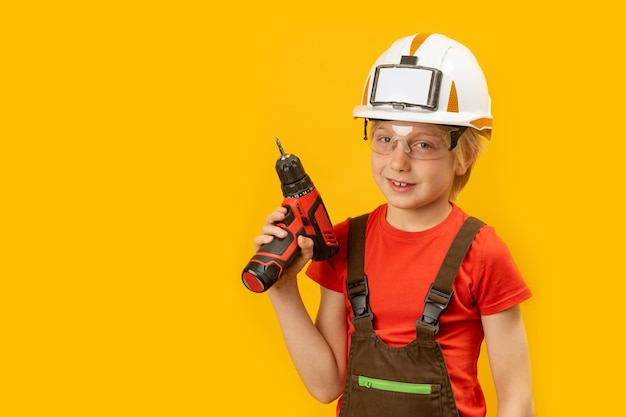 Portrait of boy as worker on yellow background Child wears protective helmet and holds drill in his hand Copy space mock up