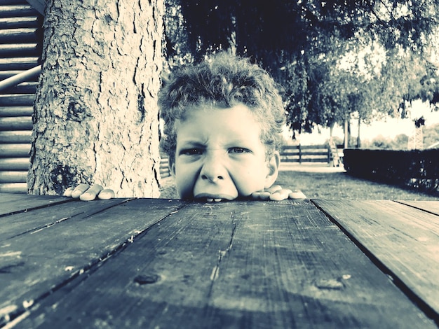 Photo portrait of boy biting wooden table outdoors