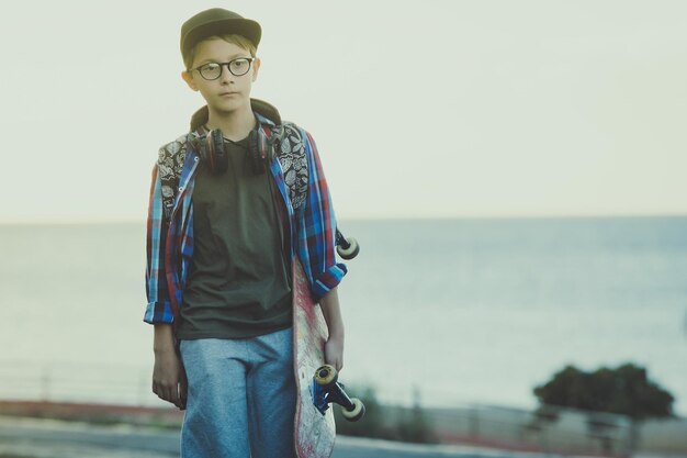 Portrait of boy holding skateboard walking on road against sea during sunset