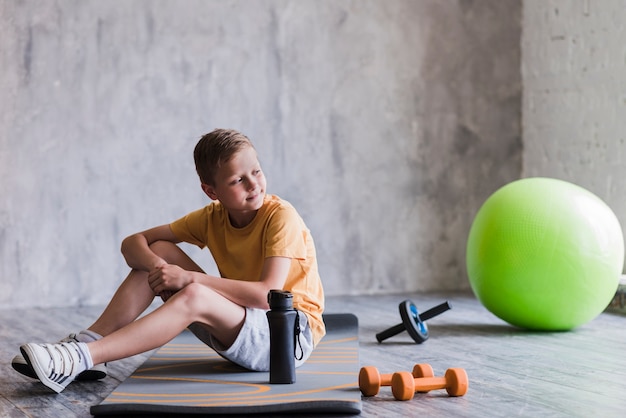 Ritratto di un ragazzo seduto vicino alla palla di pilates; manubrio; scivolo a rotelle e bottiglia d'acqua in palestra