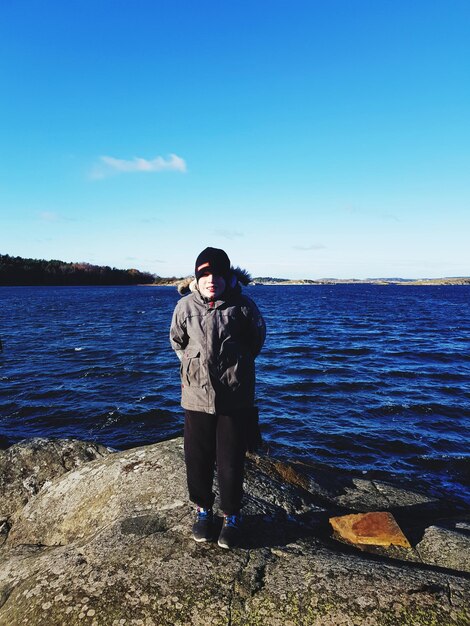 Portrait of boy standing in warm clothing on shore against blue sky