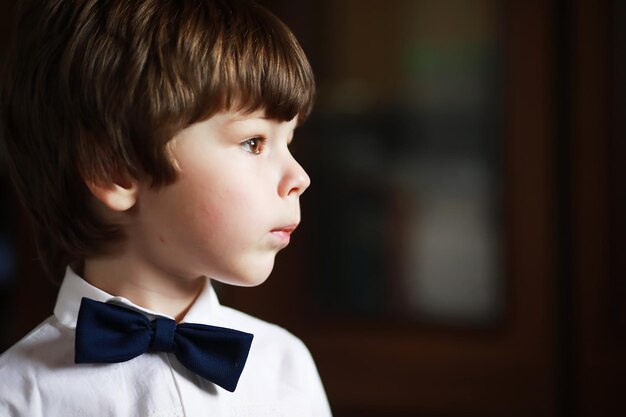 Портрет мальчика с карими глазами в рубашке и черном галстуке-бабочке на темном фоне. Ребенок эмоционален, улыбается, веселится, счастлив.