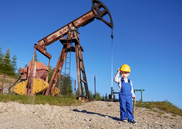 Portrait of boy in work uniform and helmet who getting acquainted with work of engineer Child holding adjustable wrench and supporting helmet with arm On the background oil well under blue sky