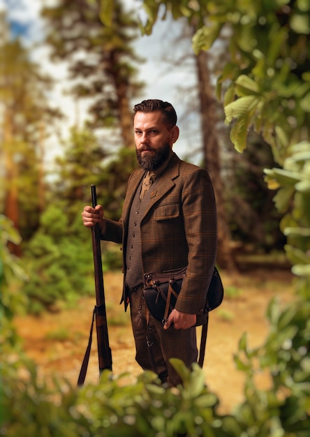 Photo portrait of breaded hunter man in traditional hunting clothing with old rifle on green forest
