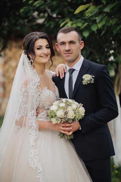 Portrait of the bride and groom in nature Stylish bride and groom in a long lace dress are hugging and posing near the trees in the garden A happy couple in love