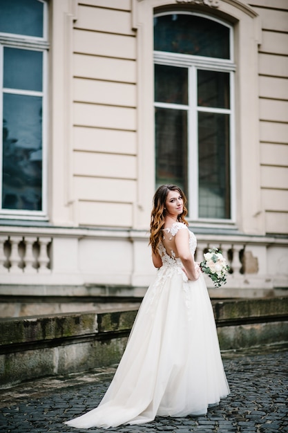 Ritratto sposa in abito da sposa con un mazzo di fiori in piedi indietro vicino al vecchio edificio esterno, palazzo d'epoca all'aperto.
