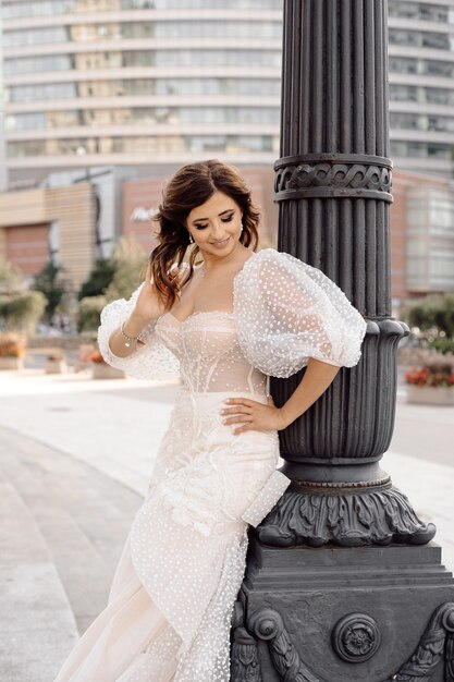 Portrait of bride with dark hair in white wedding dress at lamppost in background of city buildings, looks happily away.