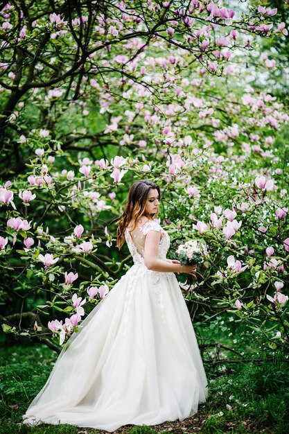 Portrait of bride with a wedding bouquet standing back, on background flowers of magnolia and greens. outdoors. Wedding location on the ceremony.