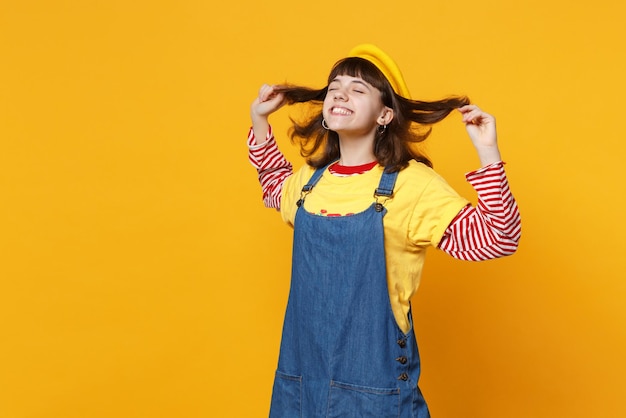 Portrait of bright girl teenager in french beret, denim sundress keeping eyes closed, holding hair isolated on yellow wall background. People sincere emotions, lifestyle concept. Mock up copy space.