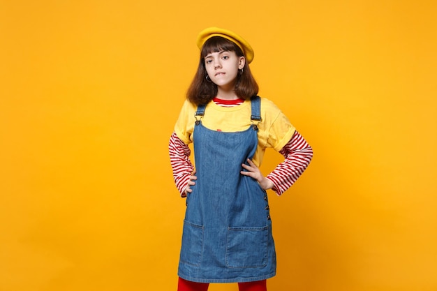 Portrait of bright girl teenager in french beret, denim sundress standing with arms akimbo on waist isolated on yellow wall background. People sincere emotions, lifestyle concept. Mock up copy space.