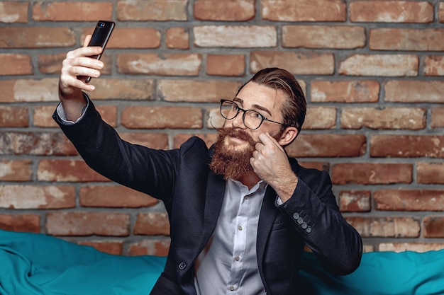 Portrait bright young gentleman with glasses, a mustache and a beard looking into a smartphone and taking a selfie