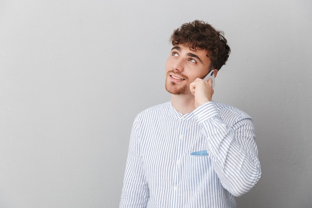 Portrait of brunette beautiful man dressed in shirt smiling while holding and talking on smartphone isolated over gray wall