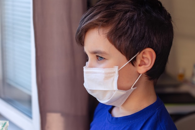 A portrait of a brunette boy in blue clothes and a white medical mask stands, looks out the window. Close up. Copy space